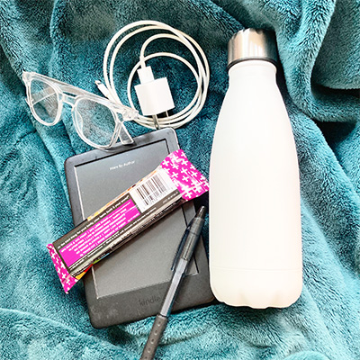 Jen's travel essentials on a blue throw - Kindle, reading glasses, phone charger, snack, white water bottle, pen